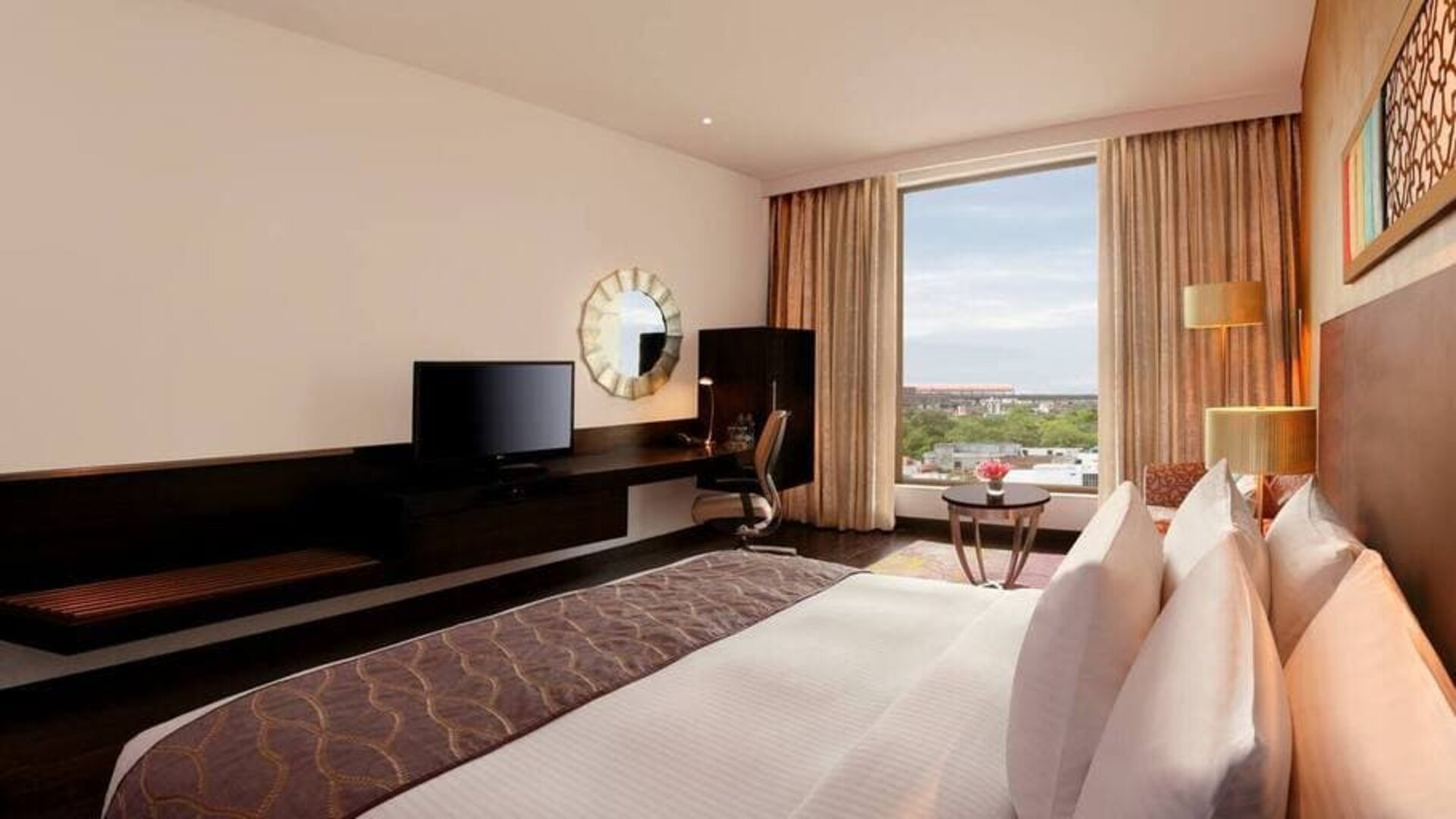 Bright king guest room with view out into Jaipur, India, complete with sitting area and long desk with television and workspace.