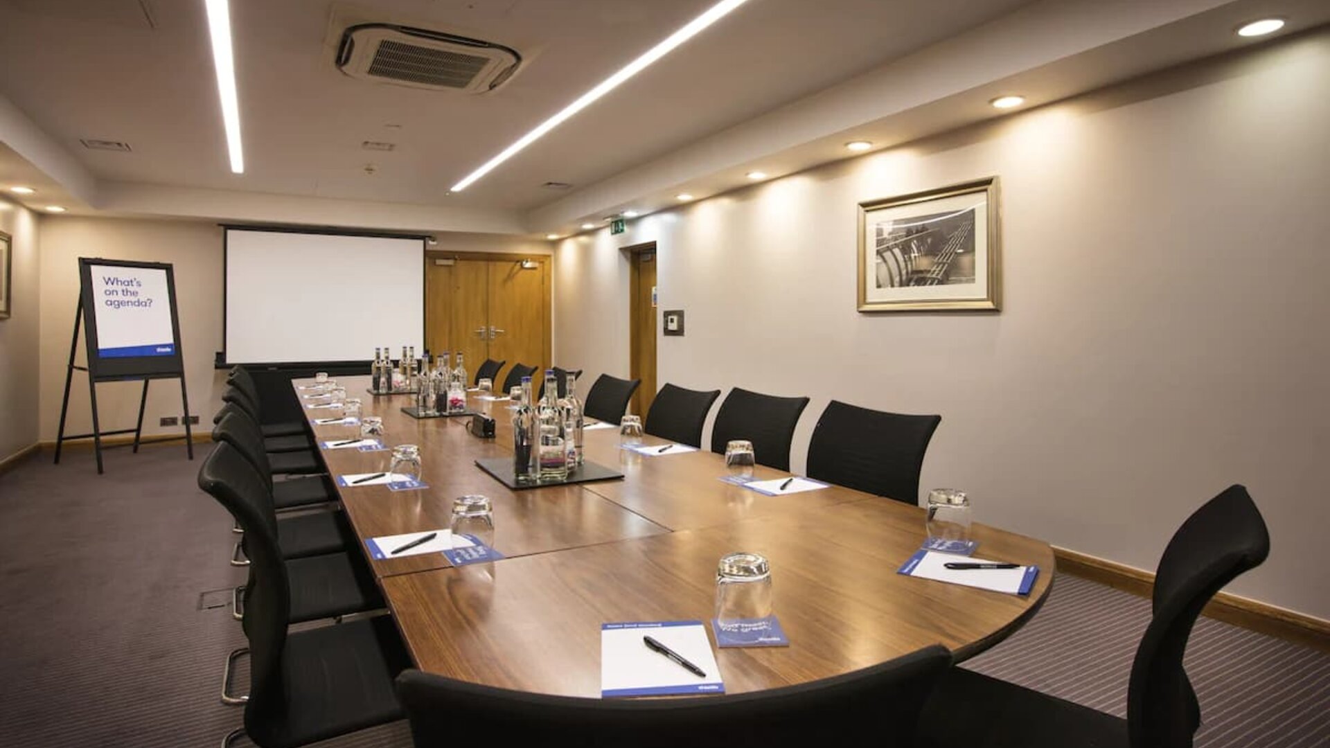 A wider shot of the meeting room with over half a dozen chairs, multiple tables, and a screen.
