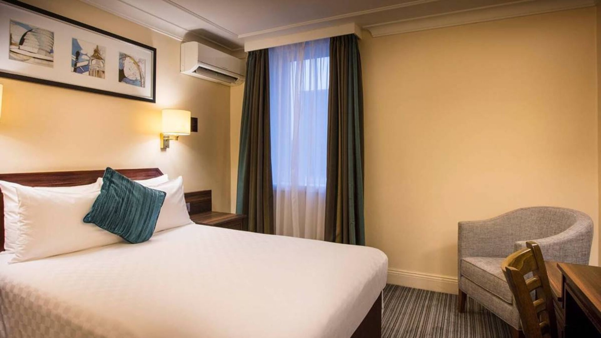 Double bed in a standard room at the Thistle London Heathrow Terminal 5, with nearby window and chair.