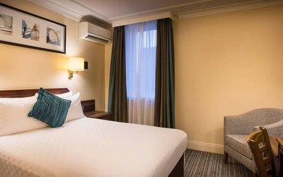 Double bed in a standard room at the Thistle London Heathrow Terminal 5, with nearby window and chair.