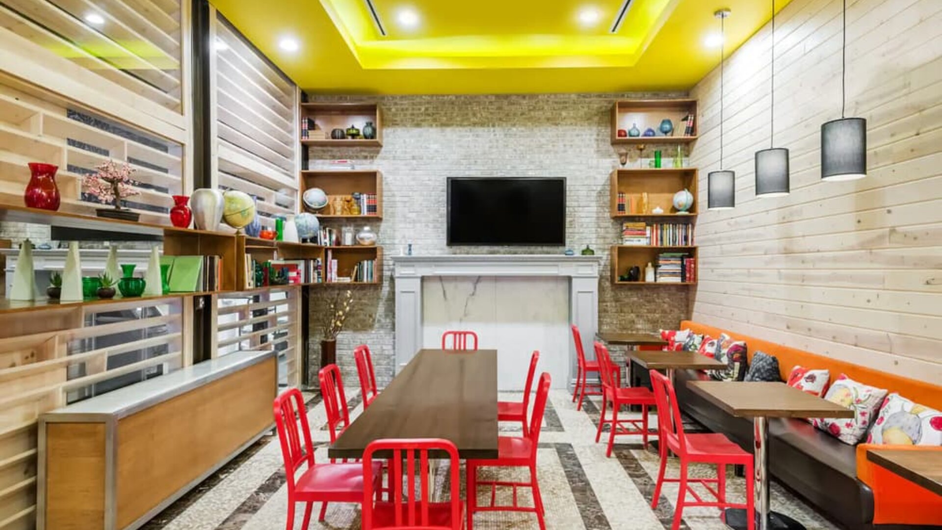 A long rectangular wooden table in the middle with red chairs. Several other smaller tables for 4 to work, eat, socialize at. Yellow lit ceiling and white fireplace mantle frames the room.