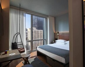 Moxy NYC Downtown double bed guest room, with large television, floor-to-ceiling window, and desk for working.