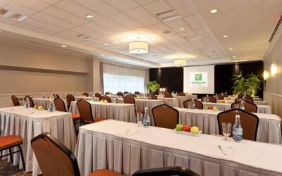 Meeting room with numerous tables and seating arranged to face a lectern, beside which is a projector screen.