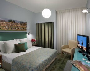 Guest bedroom with a green upholstered headboard, patterned bedspread, flat-screen TV and beige armchair at Prima Galil Tiberias.