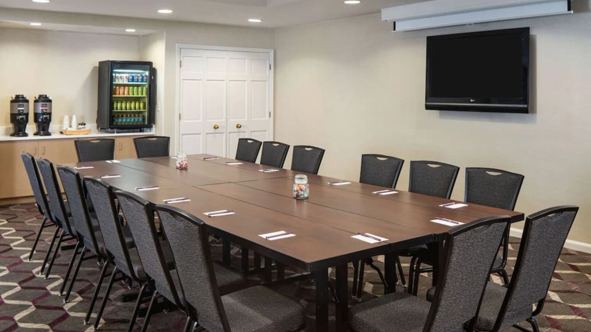 Hotel meeting room, including a long wooden table, projector screen, wall-mounted TV, and seating for over a dozen attendees.