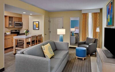 Sonesta ES Suites Cincinnati - Blue Ash guest room lounge, with armchair, sofa, and television, plus nearby kitchenette.