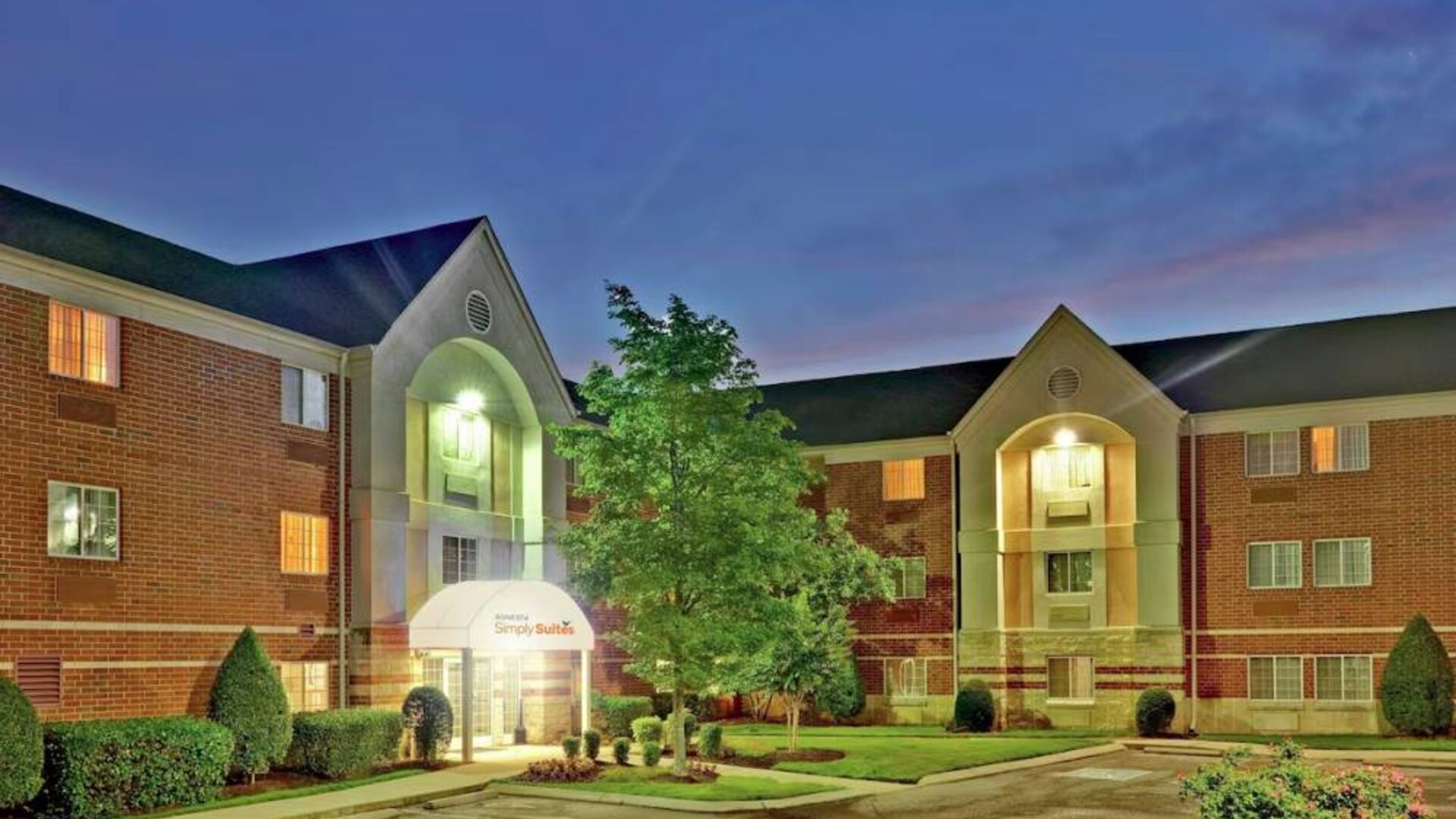 Sonesta Simply Suites Nashville Brentwood’s exterior is brightly lit and has well-tended lawns, bushes, and trees.