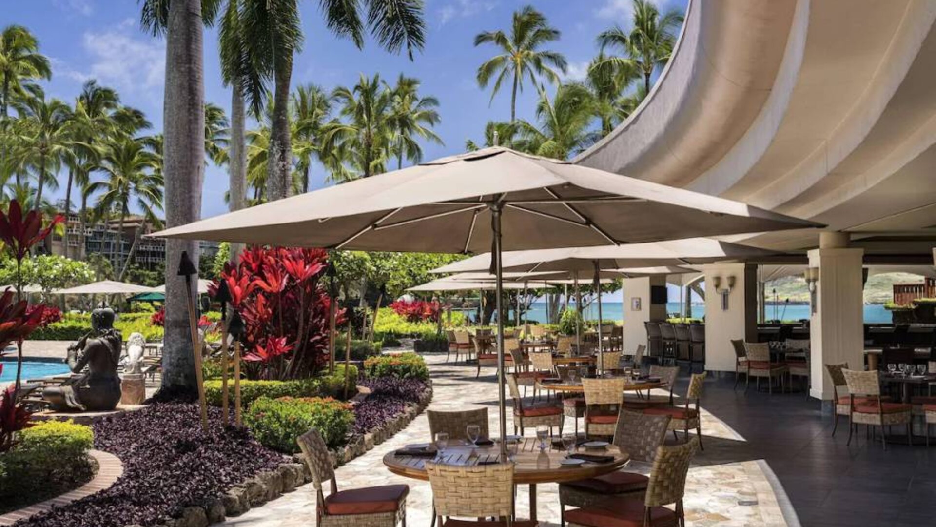The hotel’s terrace includes dining under shaded cover, with the pool and towering trees nearby.