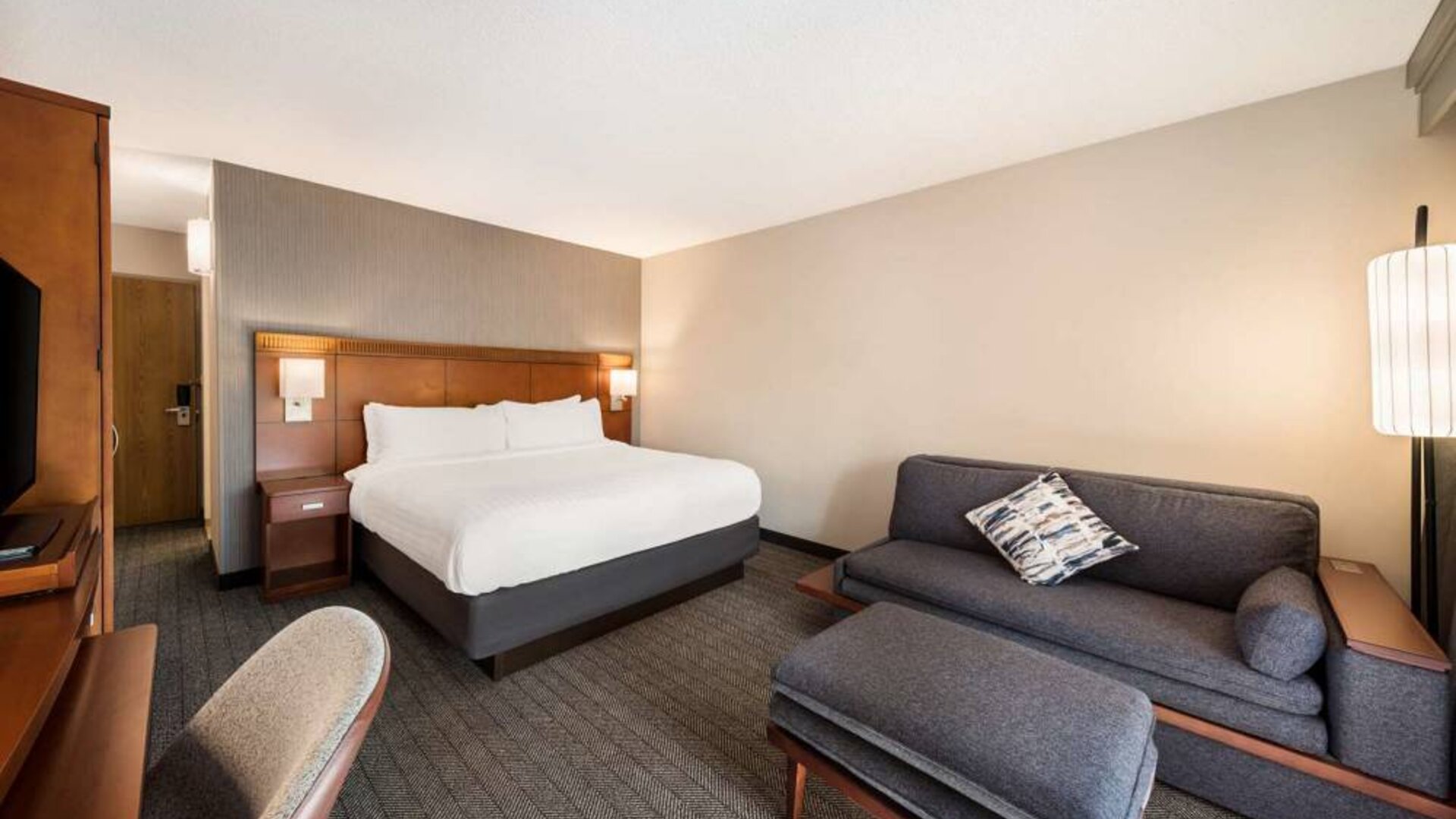 Double bed guest room in Sonesta Select San Jose Airport, furnished with sofa, chair, and TV.