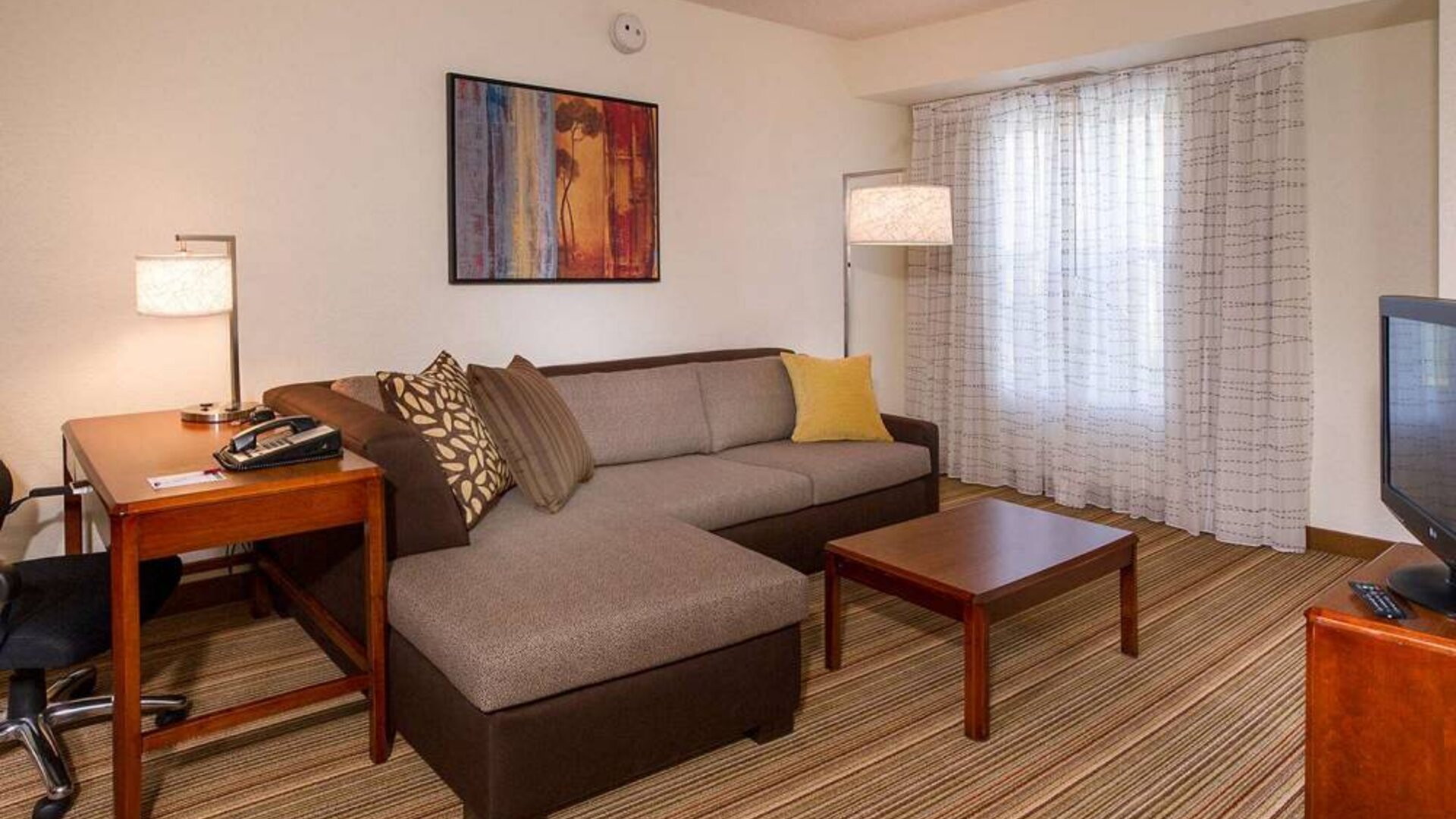 Sonesta ES Suites Charlottesville University guest room living area, including corner sofa, coffee table, TV, and window.