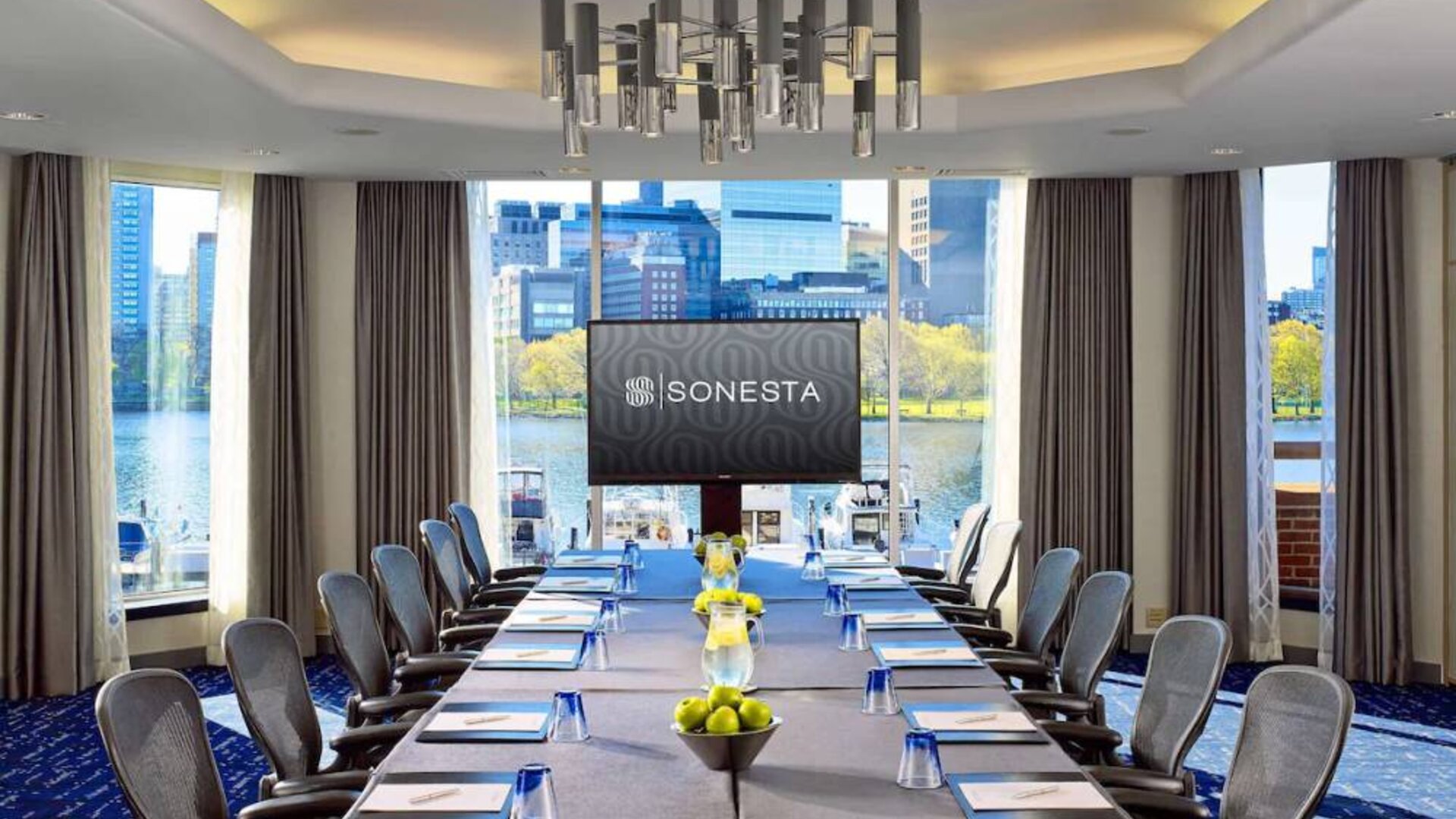 Royal Sonesta Boston meeting room, featuring large windows and high ceiling, long table and twelve surrounding chairs, plus widescreen TV.
