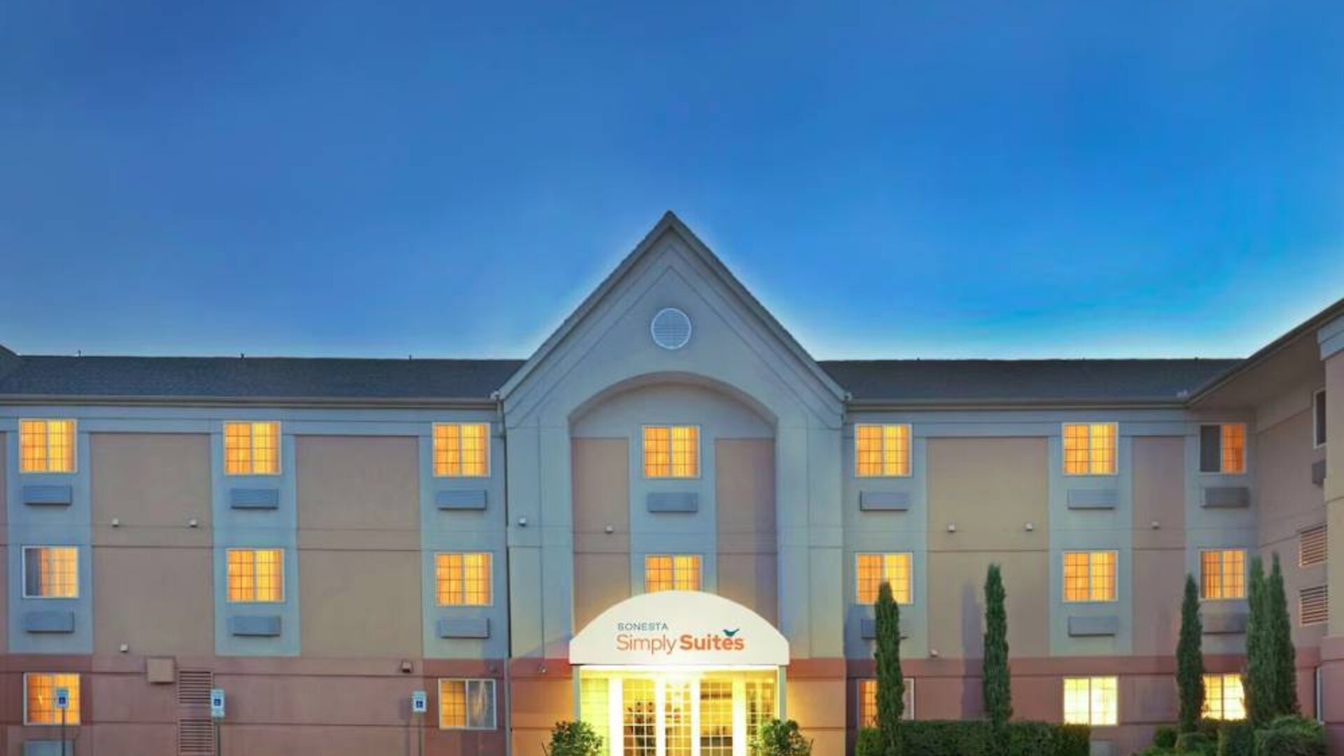 Sonesta Simply Suites Dallas Galleria’s exterior has well-kept greenery, including potted plants, clear signage, and parking spaces.