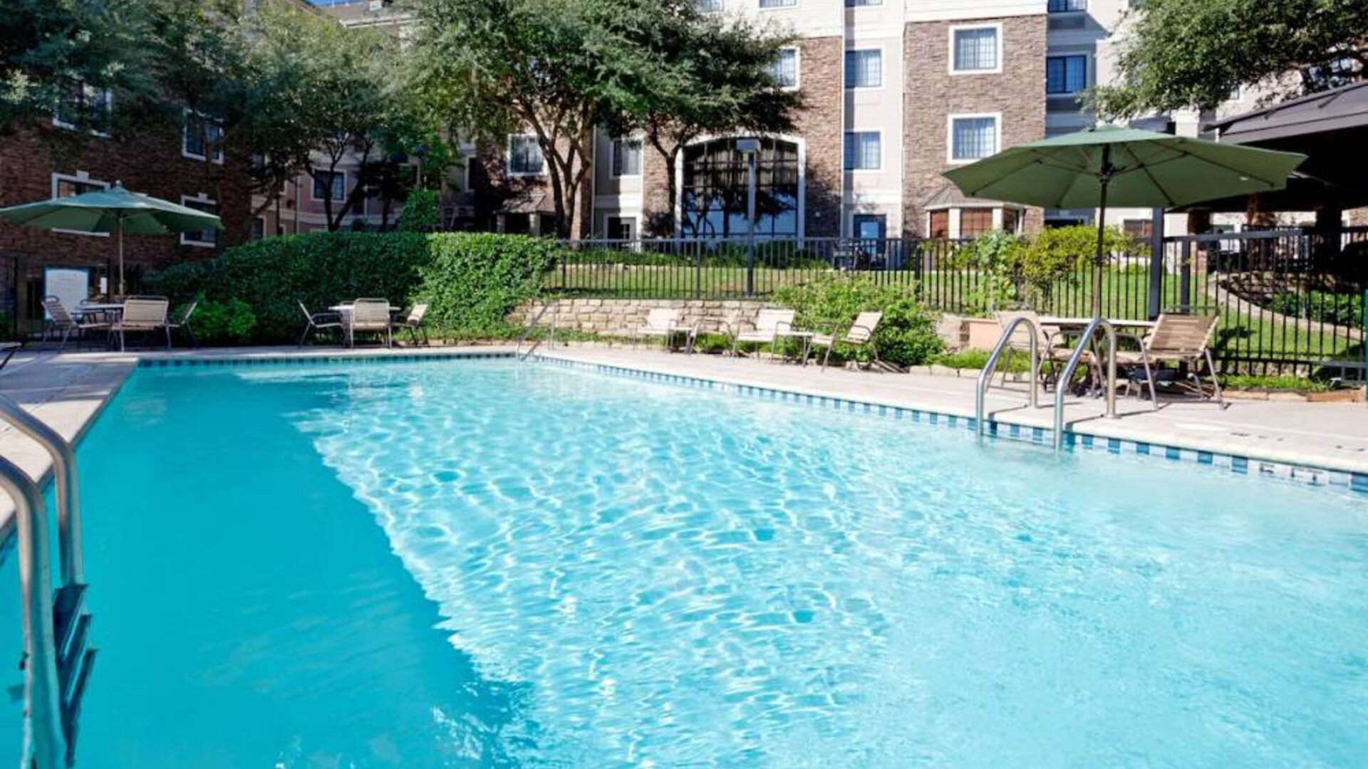 Sonesta ES Suites Austin The Domain Area’s outdoor pool features tables and chairs by the side, some of which are shaded, and nearby trees.
