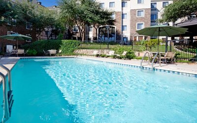 Sonesta ES Suites Austin The Domain Area’s outdoor pool features tables and chairs by the side, some of which are shaded, and nearby trees.