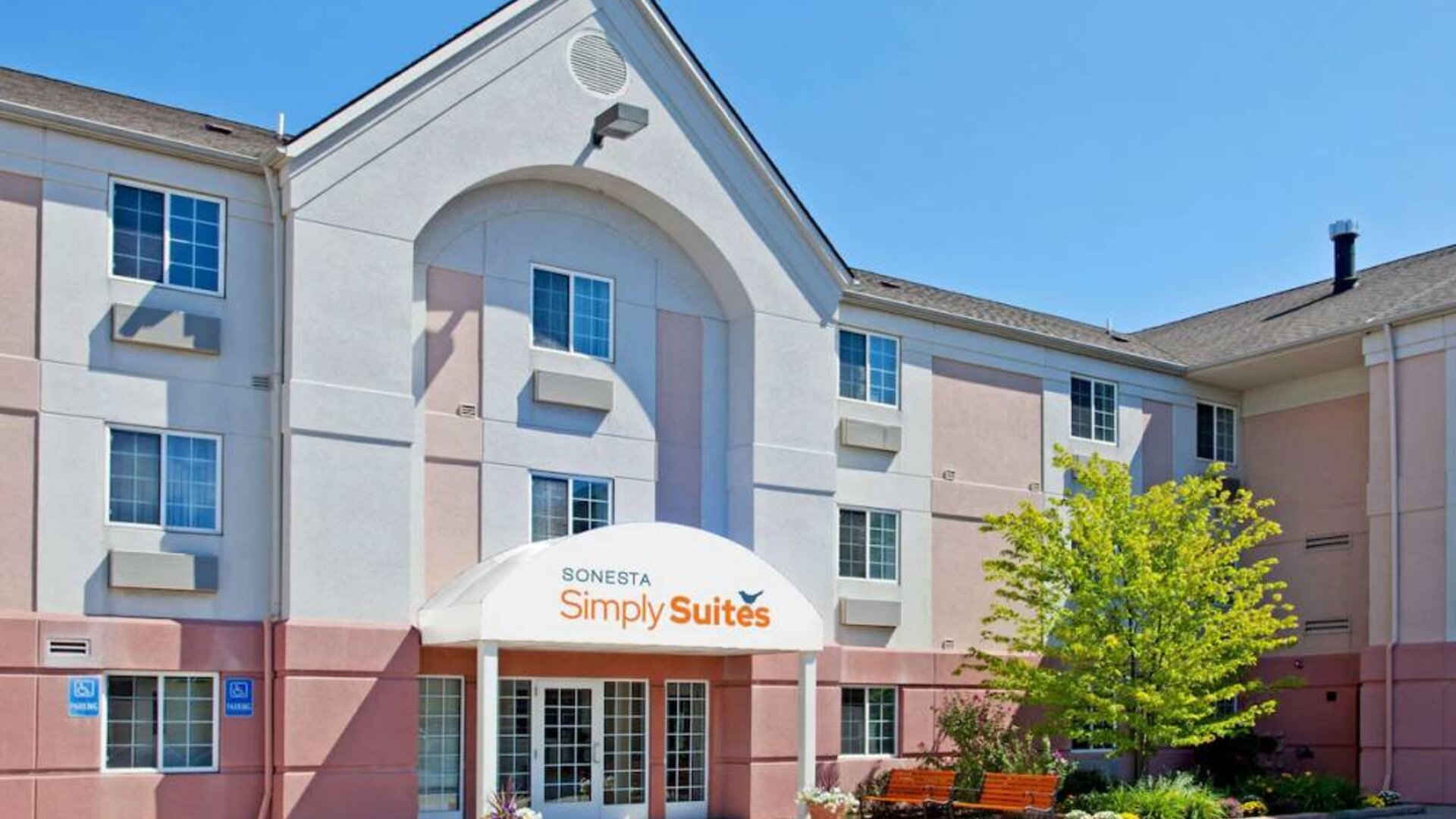 Sonesta Simply Suites Detroit Ann Arbor’s exterior features clear signage, and a pair of benches beside foliage including a tree.
