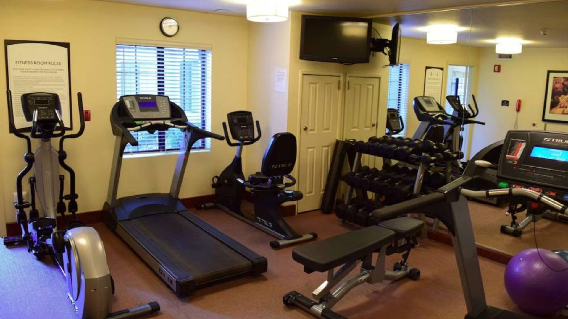 The fitness center of Sonesta ES Suites Portland Vancouver 41st Street has rows of free weights, a wall-mounted TV, and a variety of xercise machines.