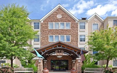 The hotel’s exterior has a covered entrance, wooden benches, and pleasant greenery including flowers and trees.