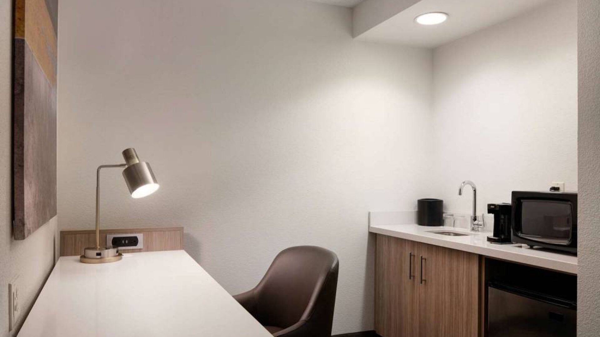 Guest room workspace in Sonesta Select Seattle Renton, including desk, chair, and lamp, with kitchenette nearby.