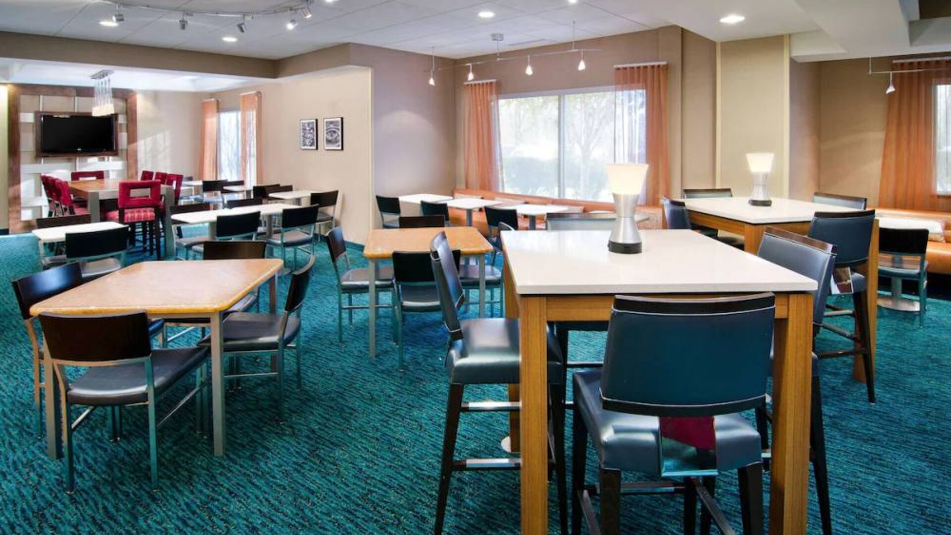 The hotel’s breakfast area is furnished with a carpet, mix of table sizes, large windows, and a TV.