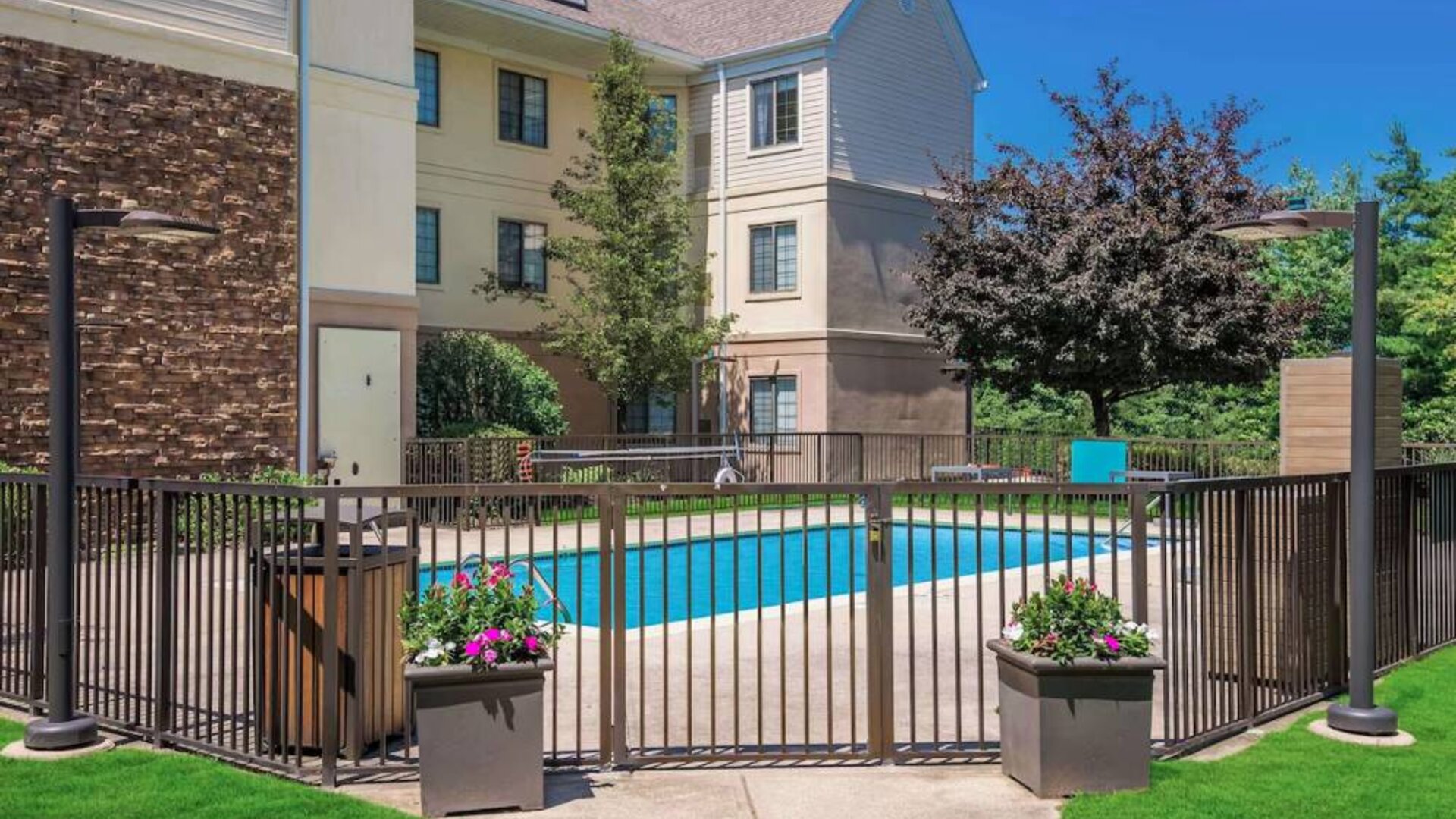 Sonesta ES Suites Andover Boston’s outdoor pool is ringed by a fence, with a backdrop of the hotel and numerous trees.