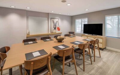 Hotel meeting room, with long wooden table and seating for nine, art and a whiteboard on the wall, and a large television. 
