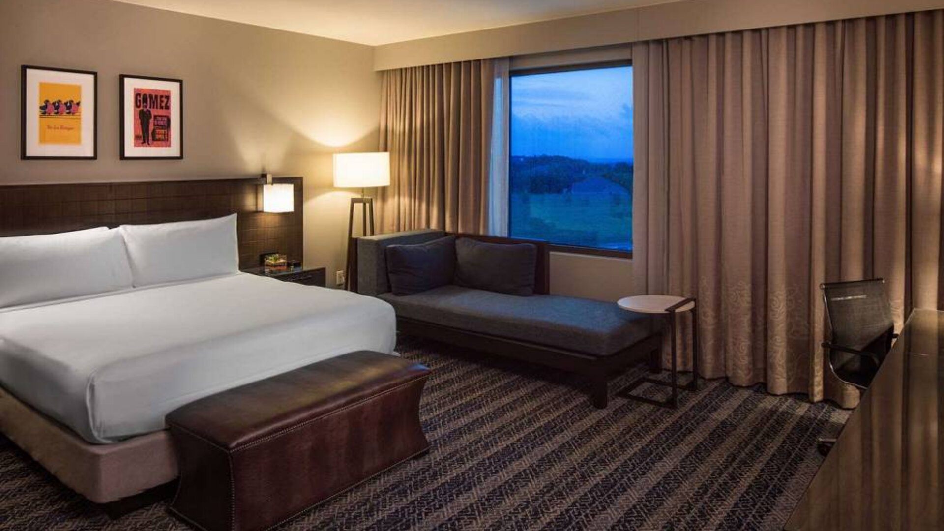 Sonesta Bee Cave Austin double bed guest room, furnished with art on the wall, desk and chair, and a sofa beside the window.