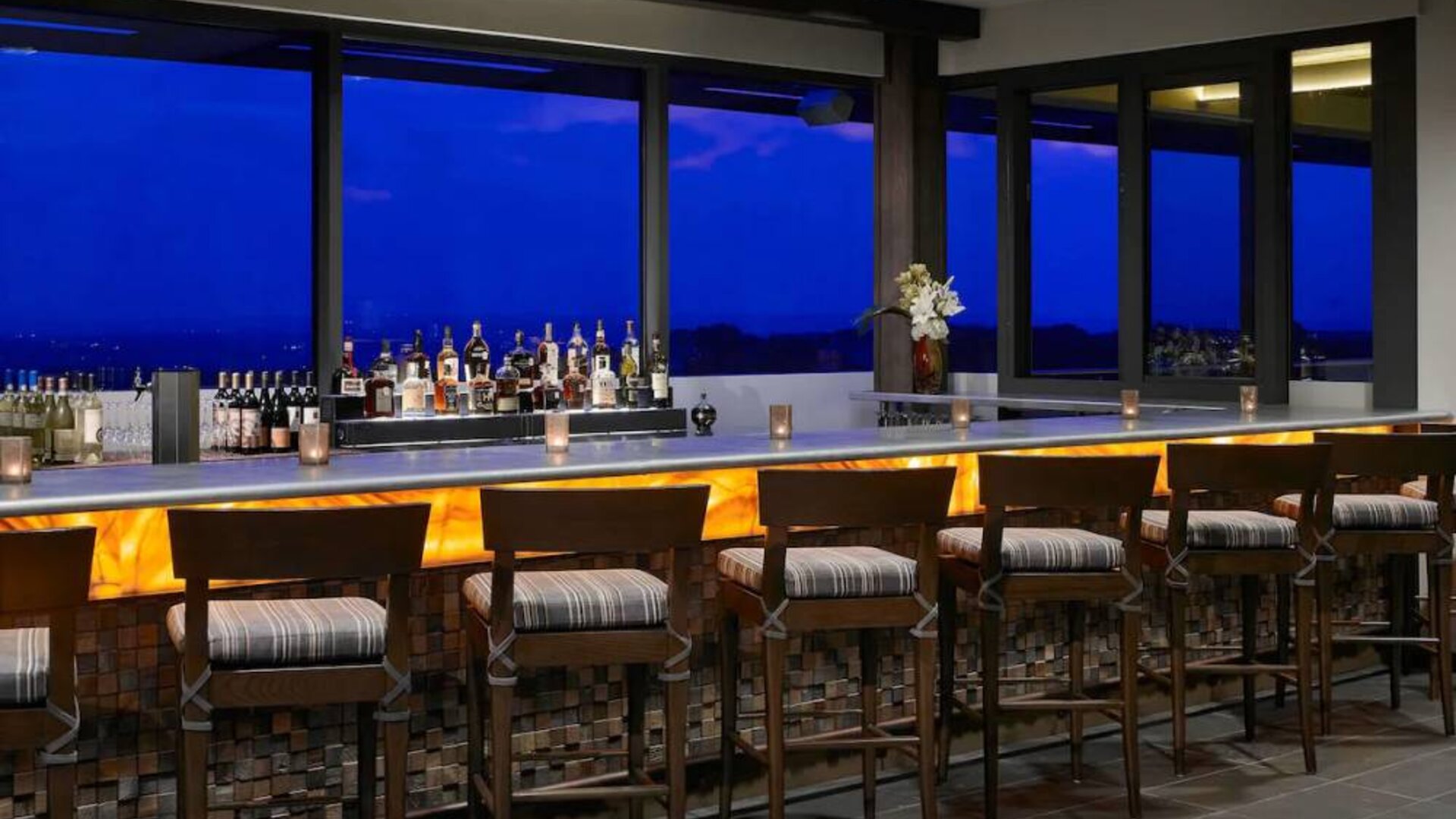 The hotel bar has traditional tall bar stools, a hard floor, a wide array of beverages, and large windows.