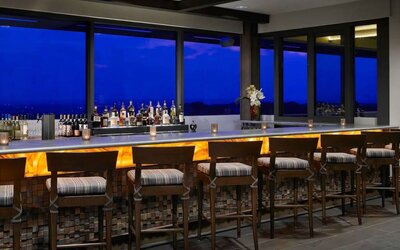 The hotel bar has traditional tall bar stools, a hard floor, a wide array of beverages, and large windows.