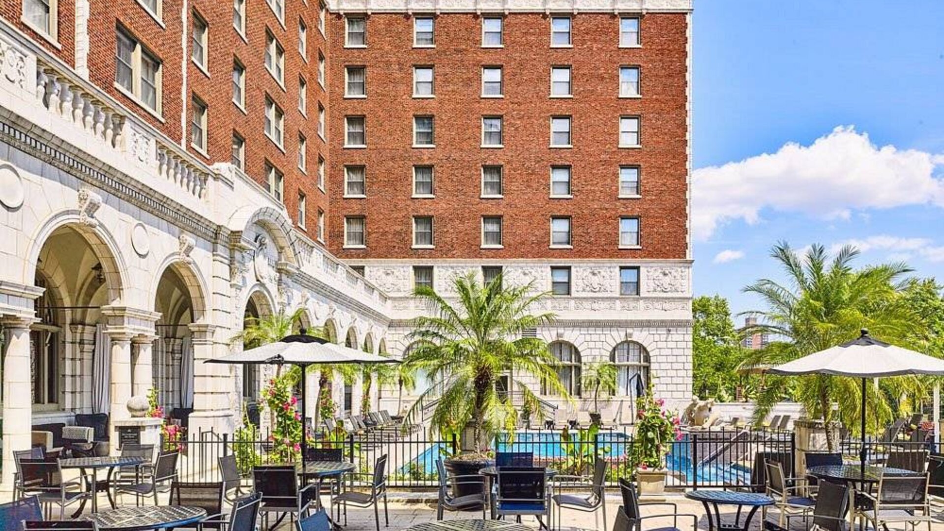 The hotel patio features tables and chairs, some shaded, overlooking the outdoor pool.