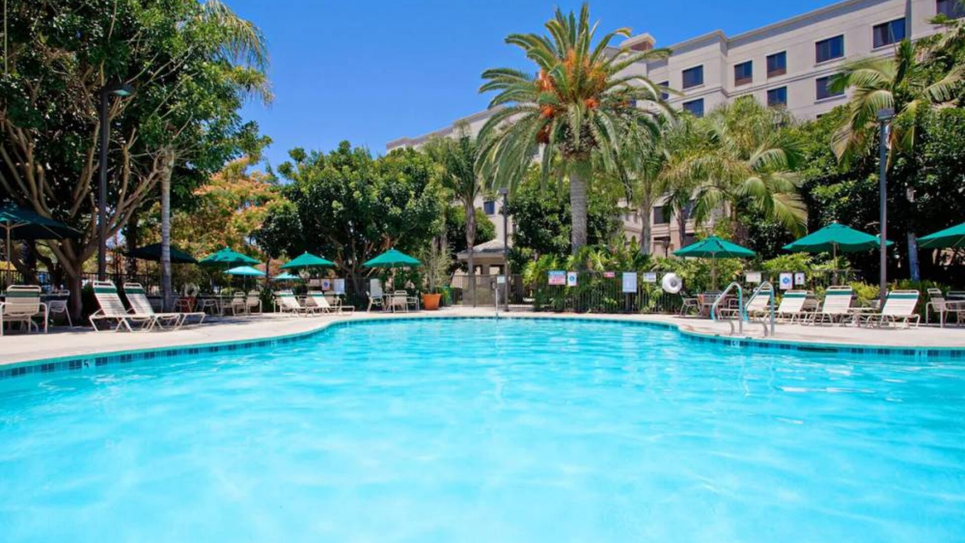 The outdoor pool at Sonesta ES Suites Anaheim Resort Area features nearby sun loungers and shaded tables and chairs, with numerous trees close by.