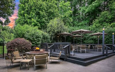 Sonesta Charlotte Executive Park’s patio includes shaded tables and chairs, and a fire pit surrounded by chairs and coffee tables, with abundant greenery nearby.