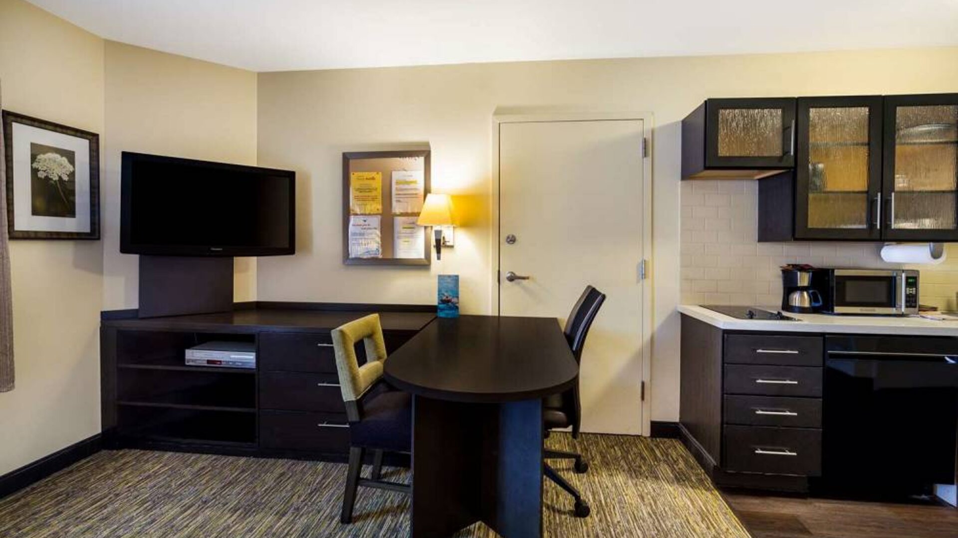 Sonesta Simply Suites St Louis Earth City guest room workspace, furnished with desk and chairs, with TV and kitchen nearby.