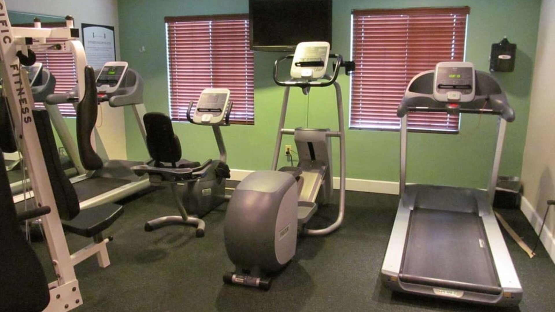 The hotel fitness center has two windows fitted with blinds, a wall-mounted TV, and  assorted exercise machines.
