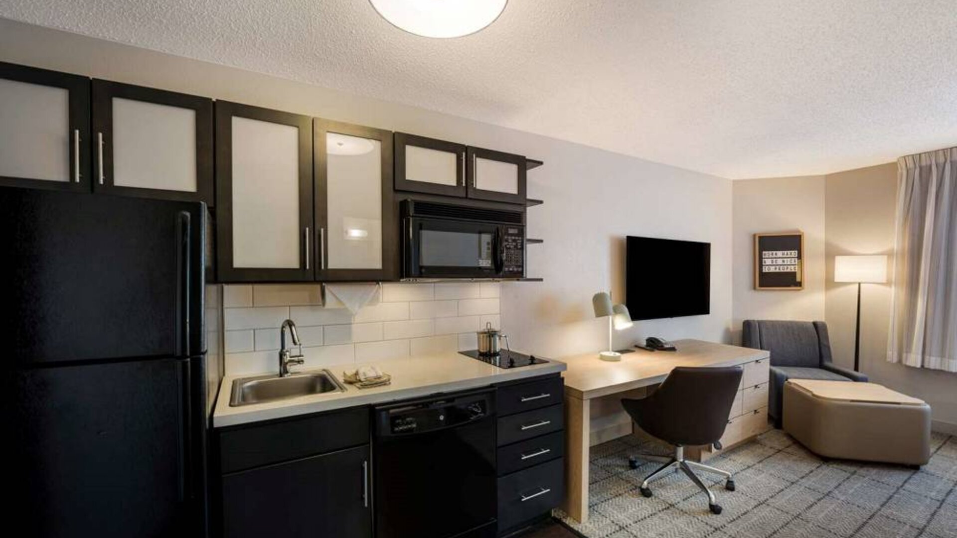 Sonesta Simply Suites Jersey City guest room kitchen, with microwave, oven, hob, and fridge-freezer, plus nearby chair and TV.