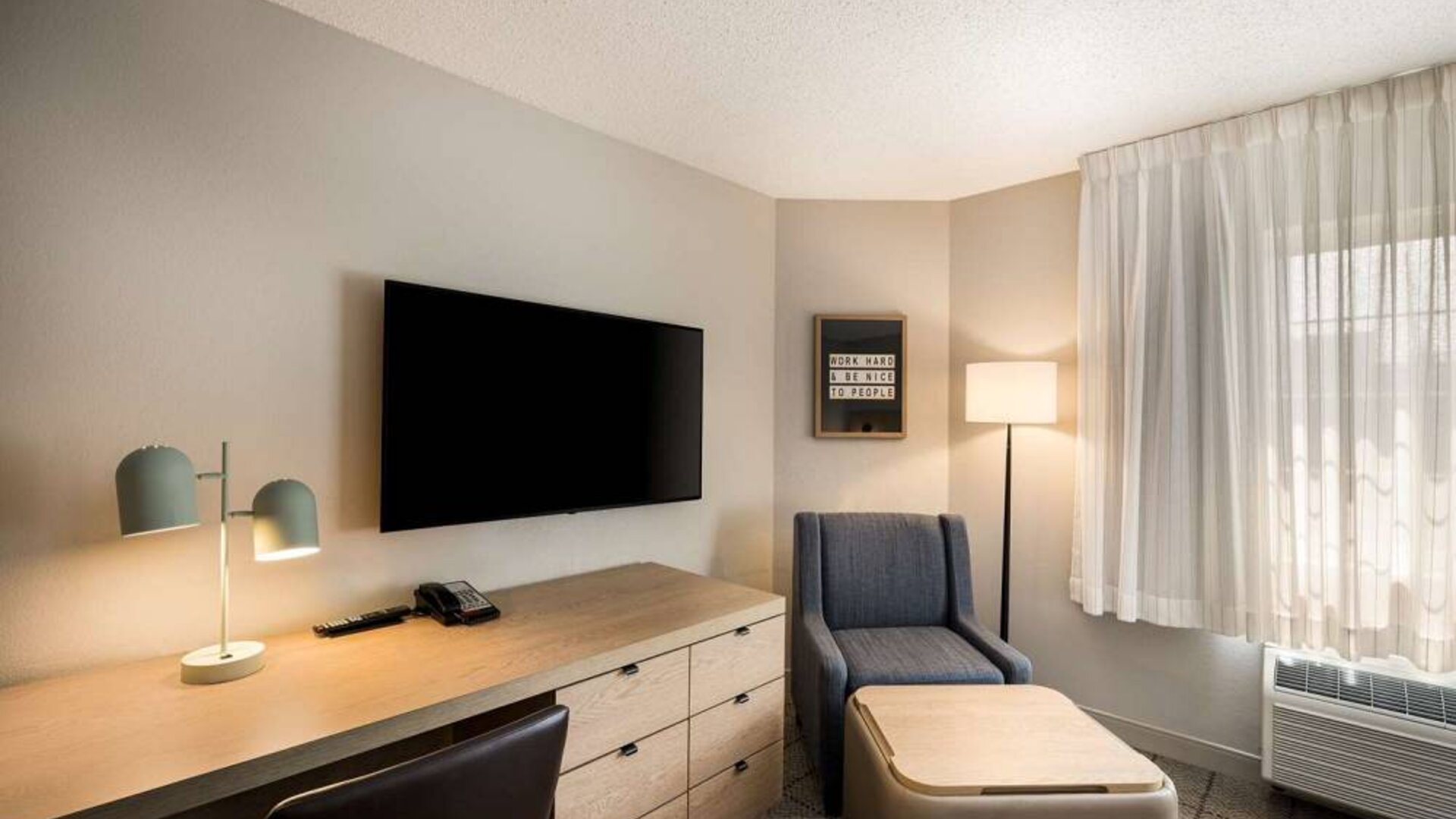 Sonesta Simply Suites Jersey City guest room workspace, including desk, chair, and lamp, with nearby TV and armchair.