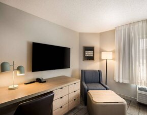 Sonesta Simply Suites Jersey City guest room workspace, including desk, chair, and lamp, with nearby TV and armchair.