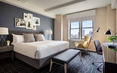 Double bed guest room in The Shelburne Sonesta New York, featuring a window, art on the wall, and a workspace desk, chair, and lamp.