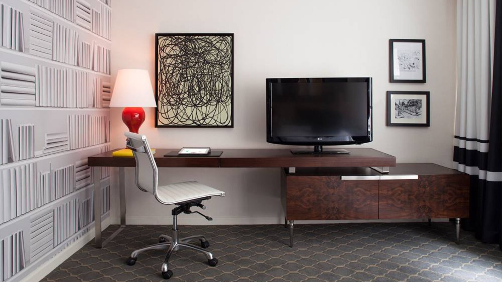 Guest room workspace in The Fifty Sonesta Select New York, featuring desk, chair, and lamp, with art on the wall and a nearby TV.