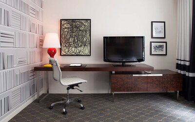 Guest room workspace in The Fifty Sonesta Select New York, featuring desk, chair, and lamp, with art on the wall and a nearby TV.