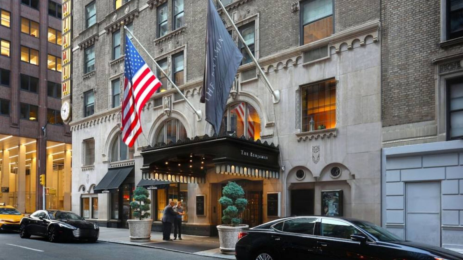 The Benjamin Royal Sonesta New York’s exterior features the hotel’s name on the awning and flag, with a Stars and Stripes flag beside it, and large potted plants.