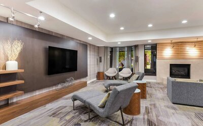 The hotel lobby includes a large, wall-mounted TV faced by a pair of reclined seats, with a fireplace and sofa nearby.