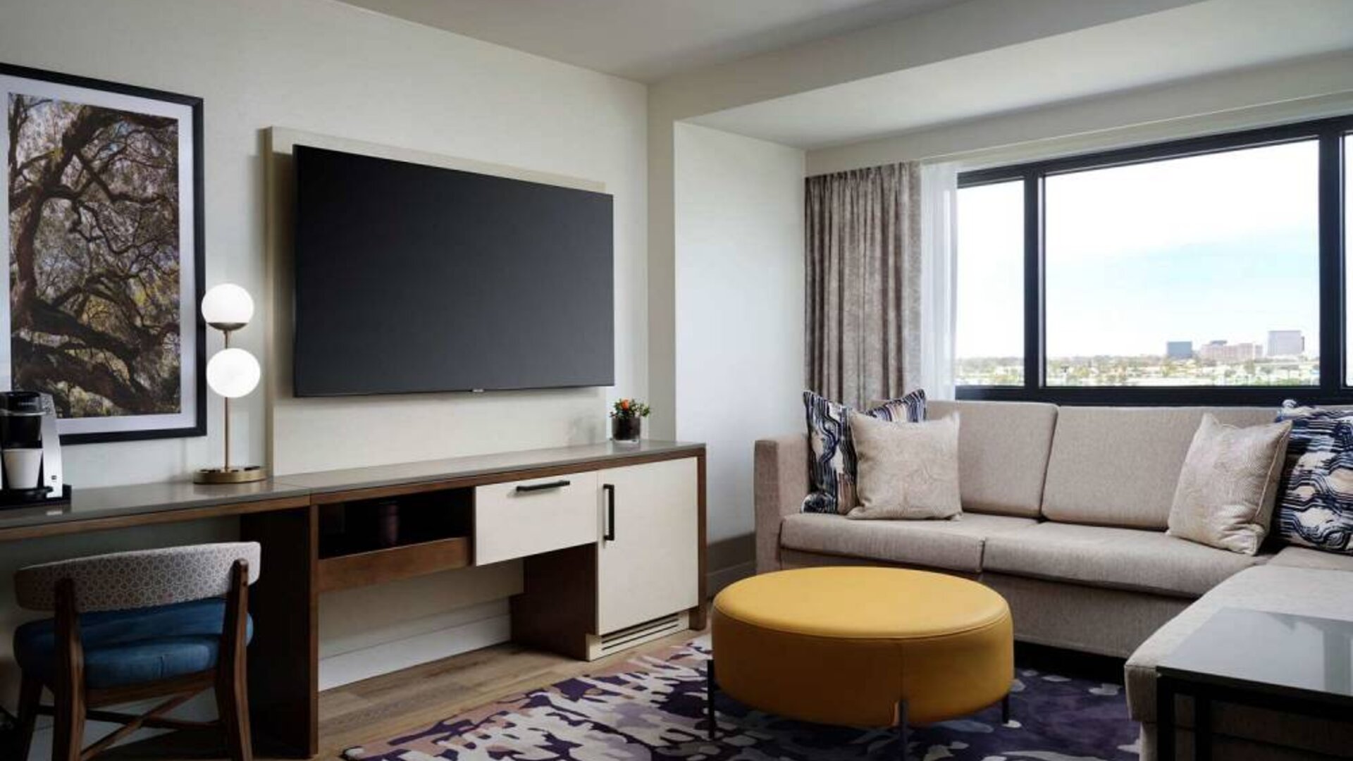 Sonesta Irvine guest room living area, including corner sofa, chair, window, and large, wall-mounted TV.