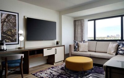Sonesta Irvine guest room living area, including corner sofa, chair, window, and large, wall-mounted TV.