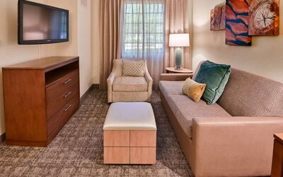 Sonesta ES Suites Fort Lauderdale Plantation guest room living area, furnished with sofa, armchair, window, and a widescreen television.