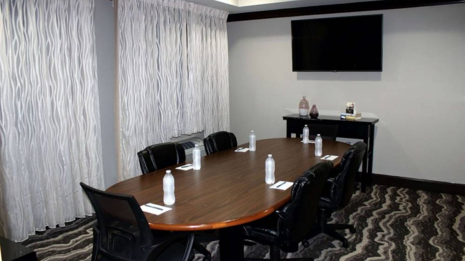 Hotel meeting room, including a wooden table and five leather chairs around it, two windows, and a large TV on the wall.
