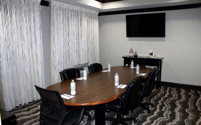 Hotel meeting room, including a wooden table and five leather chairs around it, two windows, and a large TV on the wall.