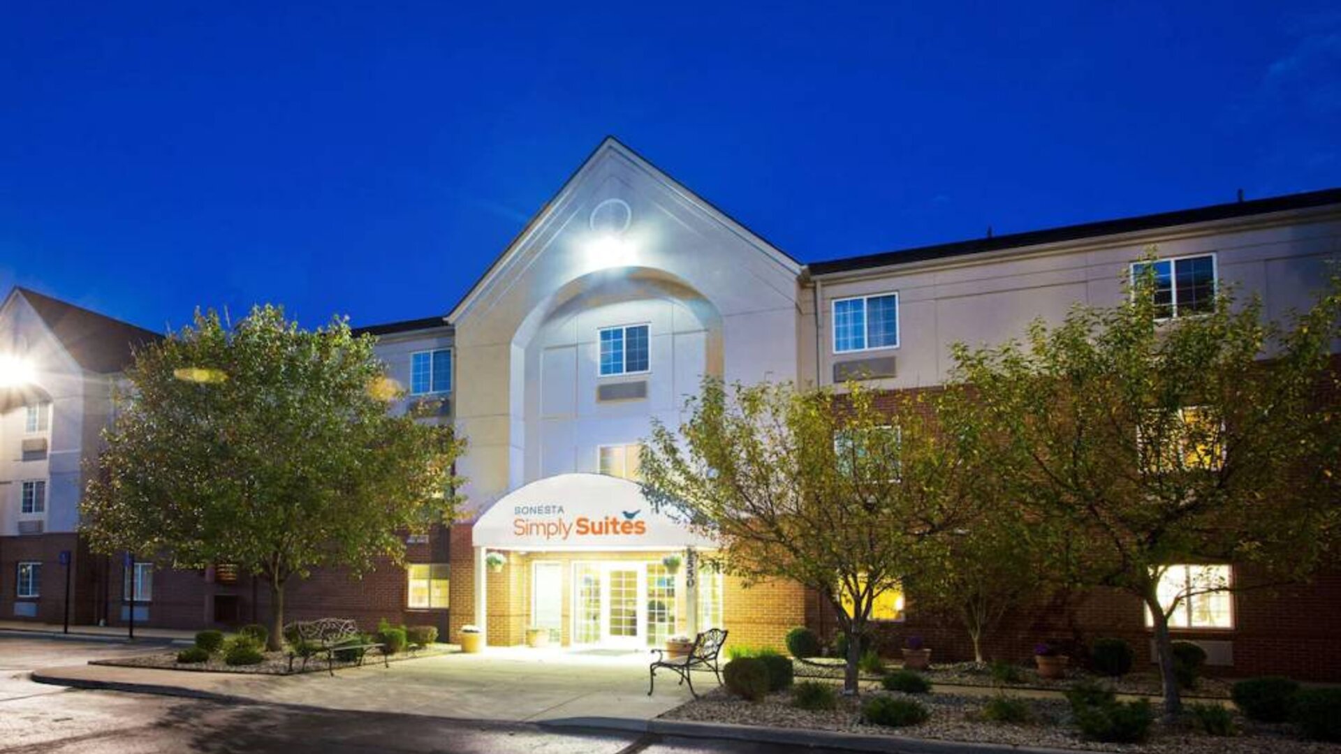 The hotel’s exterior has potted plants and benches by the front door, with trees and bushes nearby.