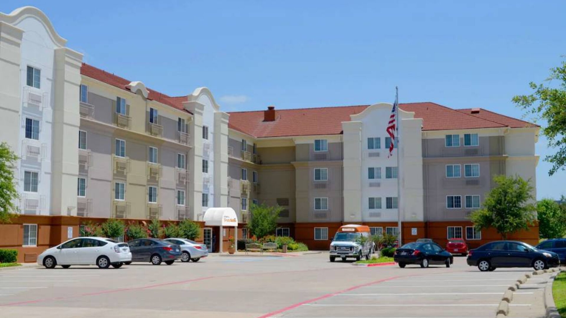 The hotel’s exterior has ample parking for guests, a US flag fluttering on a flagpole, and abundant greenery.