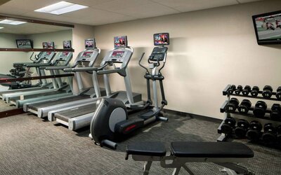 The fitness center at Sonesta Select Boca Raton comes with a wall-mounted TV, a variety of exercise machines, and rows of free weights.