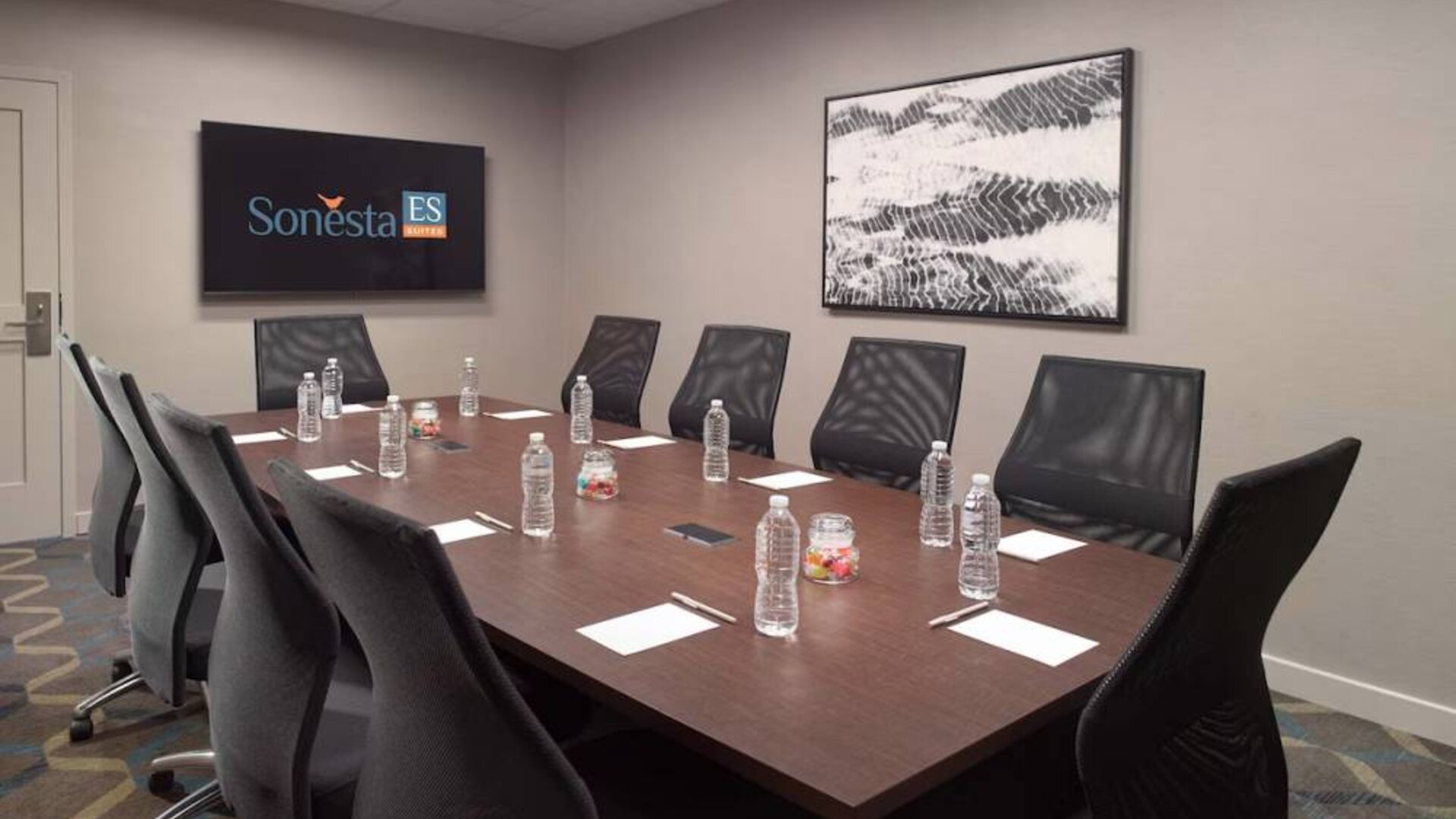 Hotel meeting room including long wooden table, 10 chairs, and a TV and art on the walls.